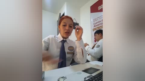 Video of a woman with light skin and red hair in a white blouse and blue tie, gesturing with her right hand, seated at a desk in a brightly lit office.