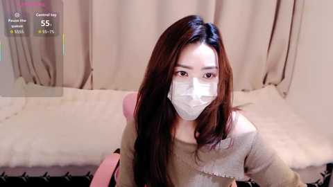 Video of an East Asian woman with long, straight brown hair, wearing a face mask, beige sweater, and pink chair, in a beige room with white bed.