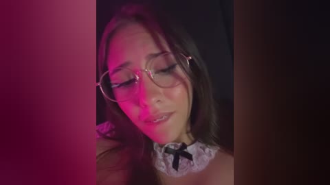 Video of a young woman with long brown hair, wearing glasses and a lace choker, under dim, pinkish-red lighting, eyes closed, slightly smiling.