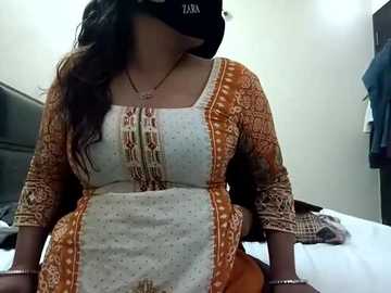 Video of a woman in a floral-patterned orange and white dress, wearing a black face mask, sitting on a bed with white sheets in a dimly lit room.