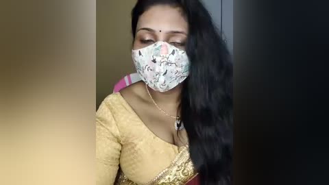 Media: Video of a South Asian woman with long black hair, wearing a yellow sari with gold embroidery, a face mask with floral pattern, and a pink headscarf, indoors.