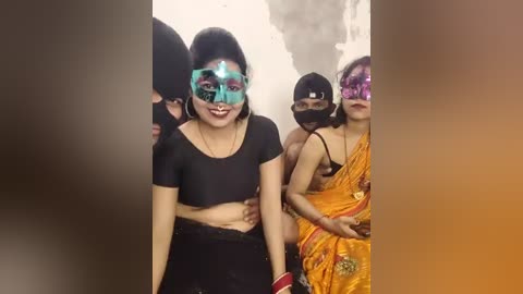 Video of a young woman in a black crop top, black skirt, and blue mask, surrounded by several women in colorful saris, all standing against a plain white wall.
