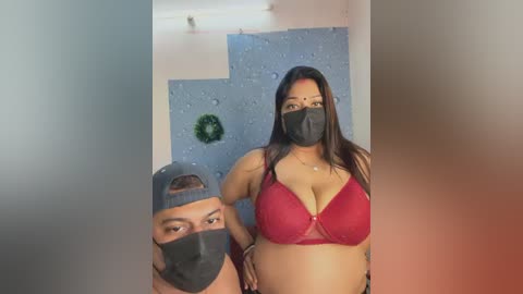 Video of two people, a man with a baseball cap and mask, and a woman with long hair, red bra, and mask, standing in a dimly lit room with blue wall art.