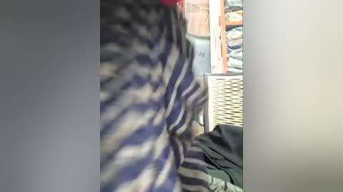 Media: A blurry, handheld video of a person wearing a striped shirt with horizontal blue and white stripes, standing in a dimly lit room with a wicker basket and clothing visible in the background.