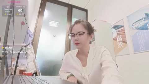 Video of a fair-skinned woman with red lipstick, glasses, and brown hair tied back, wearing a white cardigan, seated at a desk in a bright room with white walls and framed pictures.