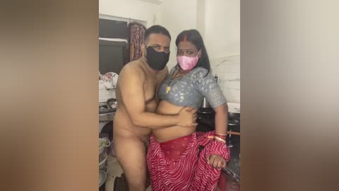 A video captures a nude man with a medium build and dark skin, wearing a black face mask, hugging a pregnant woman in a red saree with pink mask and gray top, in a cluttered, dimly lit room.