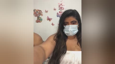 Video of a young woman with long dark hair, wearing a white top and blue mask, taking a selfie against a white wall adorned with butterfly decals.