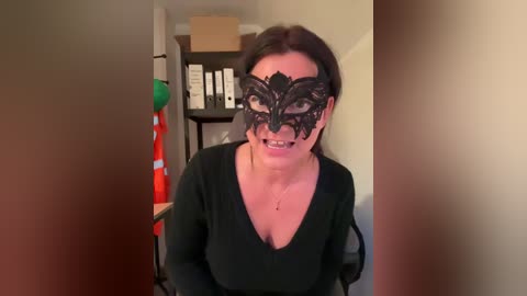 Video of a woman with a black lace masquerade mask, wearing a black long-sleeve top, sitting indoors. Background shows a cluttered room with a shelf of books and a red and green balloon.
