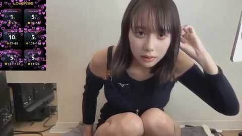 Video of a young East Asian woman with shoulder-length black hair, wearing a black off-shoulder top, sitting on a grey floor, with a TV showing a game interface in the background.