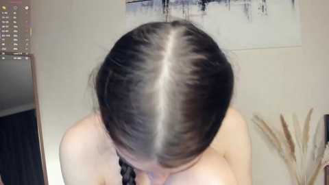 A video of a woman with long, straight black hair parted down the middle, revealing a stark white part. She has a fair skin tone. The background shows a white wall with abstract art and decorative plants.