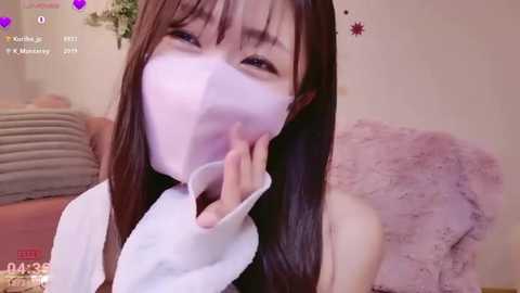 A video of an Asian woman with long brown hair wearing a white mask, smiling, in a cozy room with pink and beige decor.