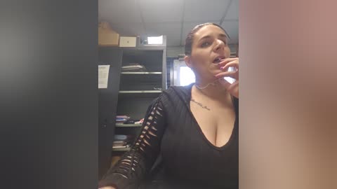 Video of a woman with fair skin and medium-length brown hair, wearing a black V-neck top with a tattoo on her chest, standing in a dimly lit office with shelves and boxes in the background.