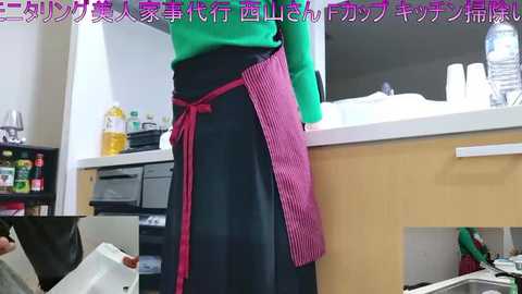 Video of a person in a green top and black skirt with a red apron, standing in a modern kitchen with white countertops, wooden cabinets, and visible appliances.