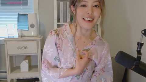 Video of a smiling Asian woman with light skin and brown hair in a floral kimono, sitting in a beige room with a white desk, bookshelf, and window with blinds.