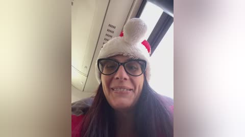 Video of a smiling woman with glasses, wearing a white Minnie Mouse hat and a red scarf, inside a bus.