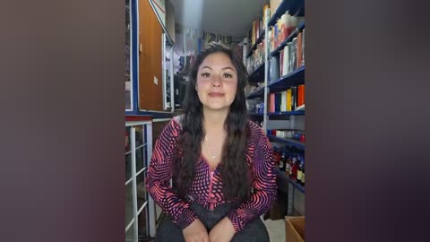 Video of a young woman with long black hair, wearing a patterned pink blouse and dark pants, sitting in a narrow, cluttered room with shelves of various items in the background.