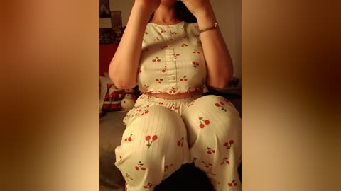 Media: Video of a light-skinned woman wearing a cherry-patterned pajama set, sitting on a bed with a red and white blanket. The background includes a framed picture and a red wall.