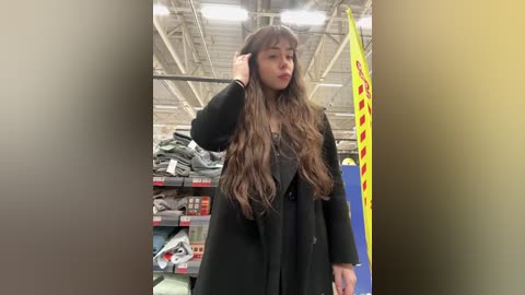 Video of a young Asian woman with long brown hair in a department store, wearing a black coat, looking confused. Background includes shelves with various items, a yellow sign, and a high ceiling.