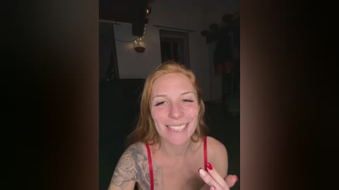 A video of a smiling, light-skinned woman with long, straight, reddish-blonde hair, wearing a red bra, with a colorful tattoo sleeve on her left arm, standing in a dimly lit, cluttered room.