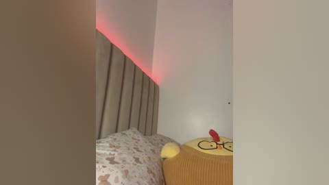 Video of a cozy bedroom corner with a beige headboard, a white wall, and a small stuffed toy featuring a yellow duck with a red beak and glasses.