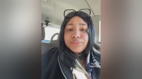 Video of a young woman with long black hair, wearing sunglasses and a black jacket, sitting in a car with a light-colored interior.