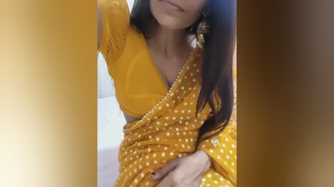 Video of a South Asian woman in a mustard-yellow sari with white polka dots, holding a phone, indoors, with a blurred background.