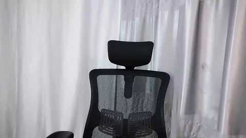 Media: Video of a black mesh office chair with a high backrest, set against a backdrop of semi-transparent white curtains. The chair's ergonomic design suggests a modern workspace.