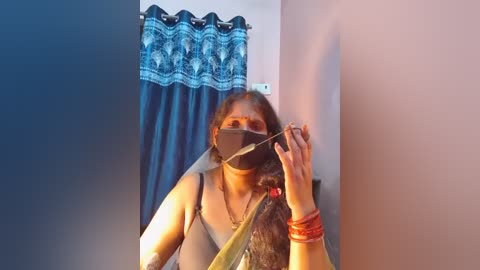 Video of a woman with long dark hair, wearing a mask, black top, and gold jewelry, applying makeup in a room with blue curtains and a pink wall.