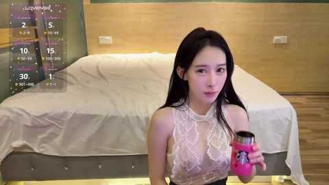 Video of an Asian woman with long black hair, wearing a sheer white lace top, holding a pink drink, in a modern bedroom with a white bed and wooden walls.