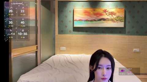Video of an Asian woman with long black hair, lying on a white bed in a modern bedroom with a green wallpaper, a framed landscape painting, and digital time display.