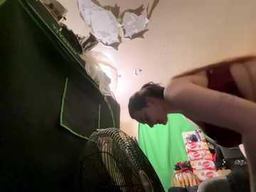 Video of a woman in a green shirt, bending over, in a cluttered, dimly lit room with peeling ceiling and green screen, revealing a wall behind.