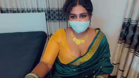 A video of a South Asian woman with medium-dark skin tone, wearing a mustard-yellow blouse, green sari with gold trim, and a blue surgical mask. She has a gold necklace and is seated on a bed with white and black striped curtains in the background.