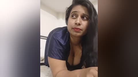 Media: A video shows a woman with long black hair, wearing a navy blue blouse, leaning forward in a domestic setting with a blurred background.