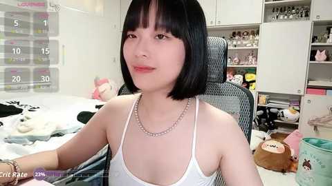 Video of a young Asian woman with straight black hair, wearing a white spaghetti-strap top and pearl necklace, sitting in a cluttered room with a calendar, plush toys, and shelves filled with knick-knacks.