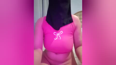 Video of a woman with a large black head covering, wearing a bright pink crop top with a white bow design. Background features a blurred bedroom setting with pink lighting.