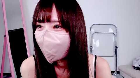 Video of an East Asian woman with long, straight, dark brown hair, wearing a light pink face mask and a black tank top, standing indoors with a white wall and a metal folding chair in the background.