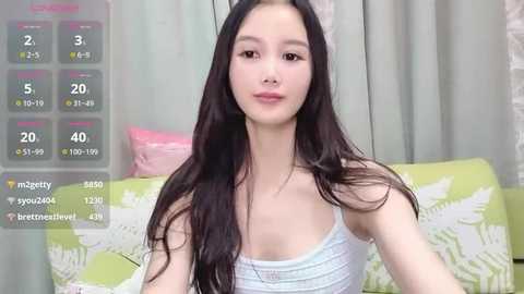 Video of a young Asian woman with long black hair, fair skin, wearing a white tank top, sitting on a green floral-patterned couch. Background features light green curtains.