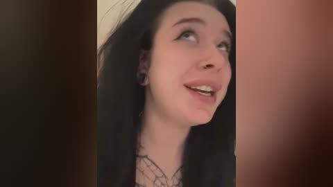 Media: Video of a young Caucasian woman with long black hair and fair skin, wearing a black lace top, smiling, with blurred, dark-haired background.