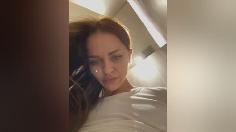A video of a young woman with fair skin and straight, shoulder-length brown hair, wearing a white t-shirt, looking downcast with a pensive expression. The background features a dimly lit, plain white wall.