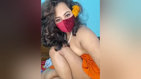 Video of a woman with curly brown hair, wearing a maroon face mask and an orange sari, sitting on a bed, with a blue wall and colorful fabric in the background.