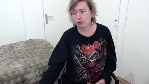 A video of a young, fair-skinned man with shoulder-length, tousled blonde hair wearing a black, graphic t-shirt featuring a skull and a red background. He sits on a black chair in a simple, white-walled room with a grey quilted blanket on the bed behind him.