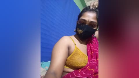 Video of a South Asian woman with medium skin tone, wearing a mustard yellow lace bra and a red saree with gold embroidery, black face mask, blue wall background, blurred.