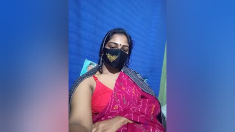Video of a South Asian woman with medium skin tone, wearing a red sari with intricate embroidery, black mask, and gray cardigan. Background features a blue wall and indistinct objects.