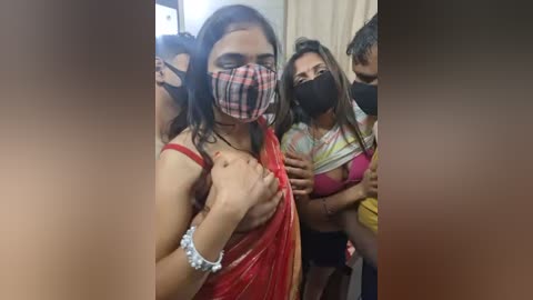 Video of four South Asian women in a crowded indoor setting, wearing face masks and colorful, revealing attire; one woman covers her chest with a red sari.