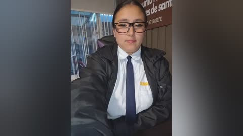 Video of a young, light-skinned Latina woman with dark hair in a bun, wearing glasses, a white shirt, dark tie, and a black jacket. She's sitting indoors, behind a counter with a sign in Spanish.