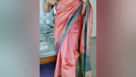 A video of a woman wearing a vibrant pink sari with green border, adjusting her sari in a bedroom with a grey bed and floral-patterned sheets.