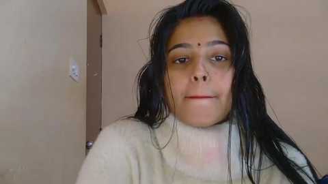Video of a young South Asian woman with long black hair, wearing a white turtleneck sweater, in a beige room with a door and light switch.