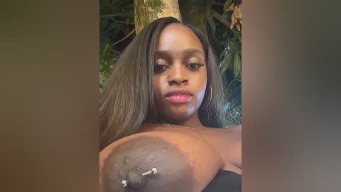 A video of a young Black woman with medium-brown skin, long straight hair, and a large areola piercing, standing in a lush, green jungle setting.