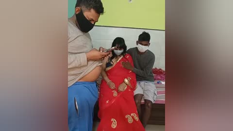 Video of three people in a hospital room: a man in a gray shirt and blue pants, a woman in a red sari, and a man in a gray shirt and white mask.