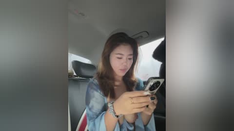 A video of an East Asian woman with long, straight brown hair, wearing a tie-dye blue shirt, sitting in a car, focused on her smartphone.